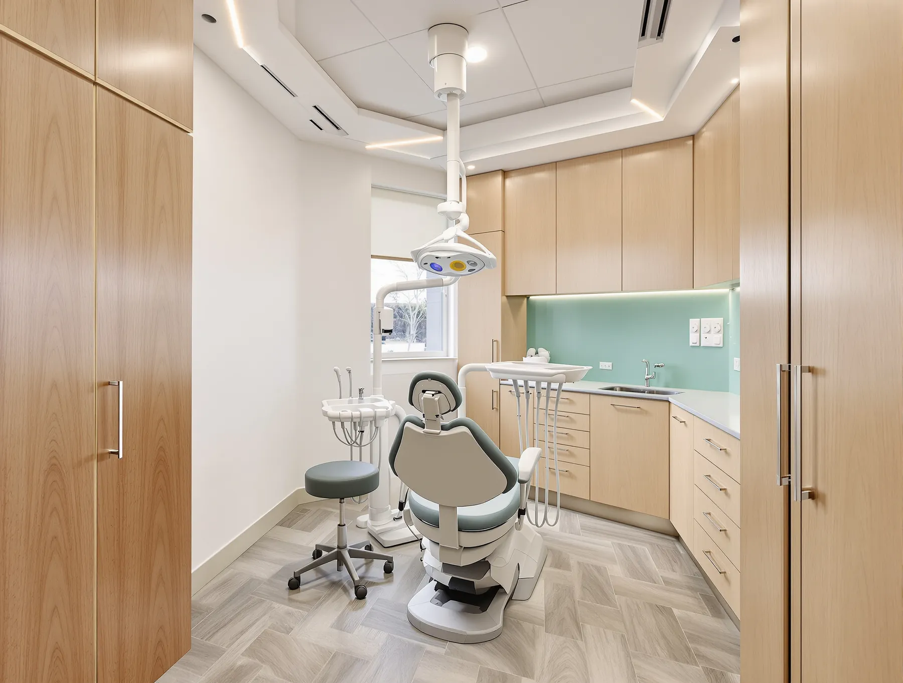 A dental surgery room with a treatment chair, timber joinery, a stainless basin and a teal splashback.