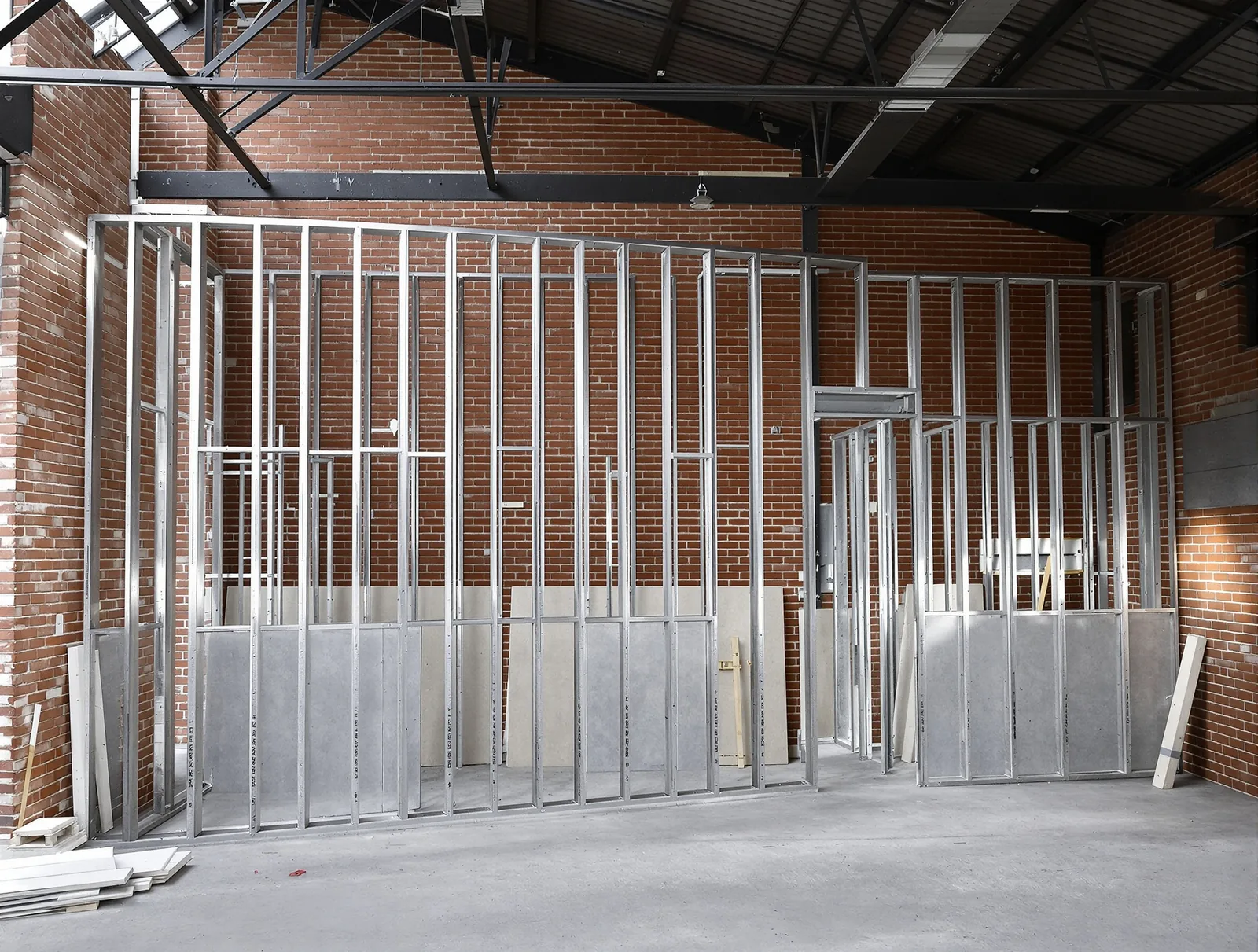 New steel stud framing standing against an exposed brick wall inside a converted warehouse, with the old steel roof left open above.