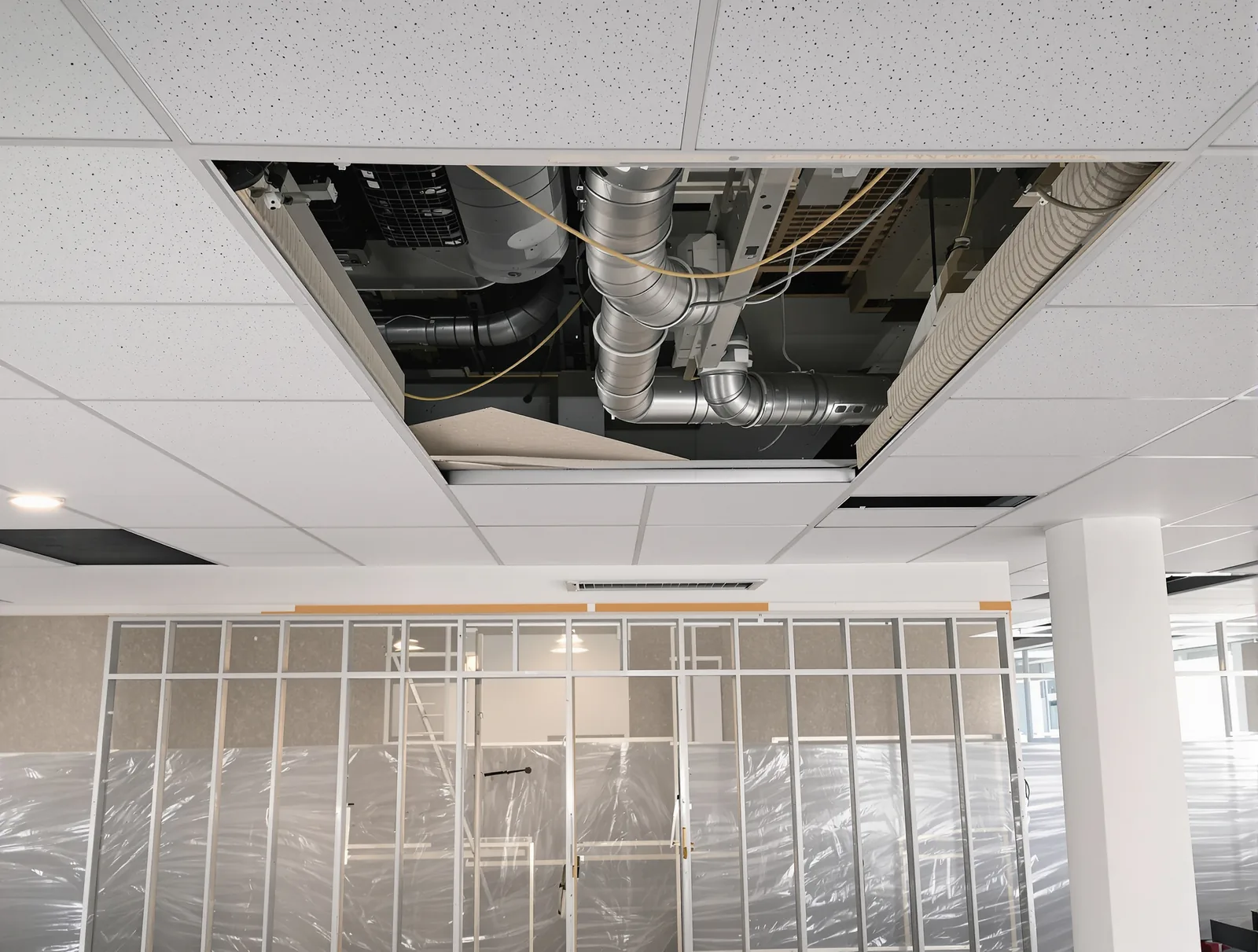 A suspended ceiling opened up with tiles stacked aside, exposing ductwork above new steel stud framing inside an occupied office.