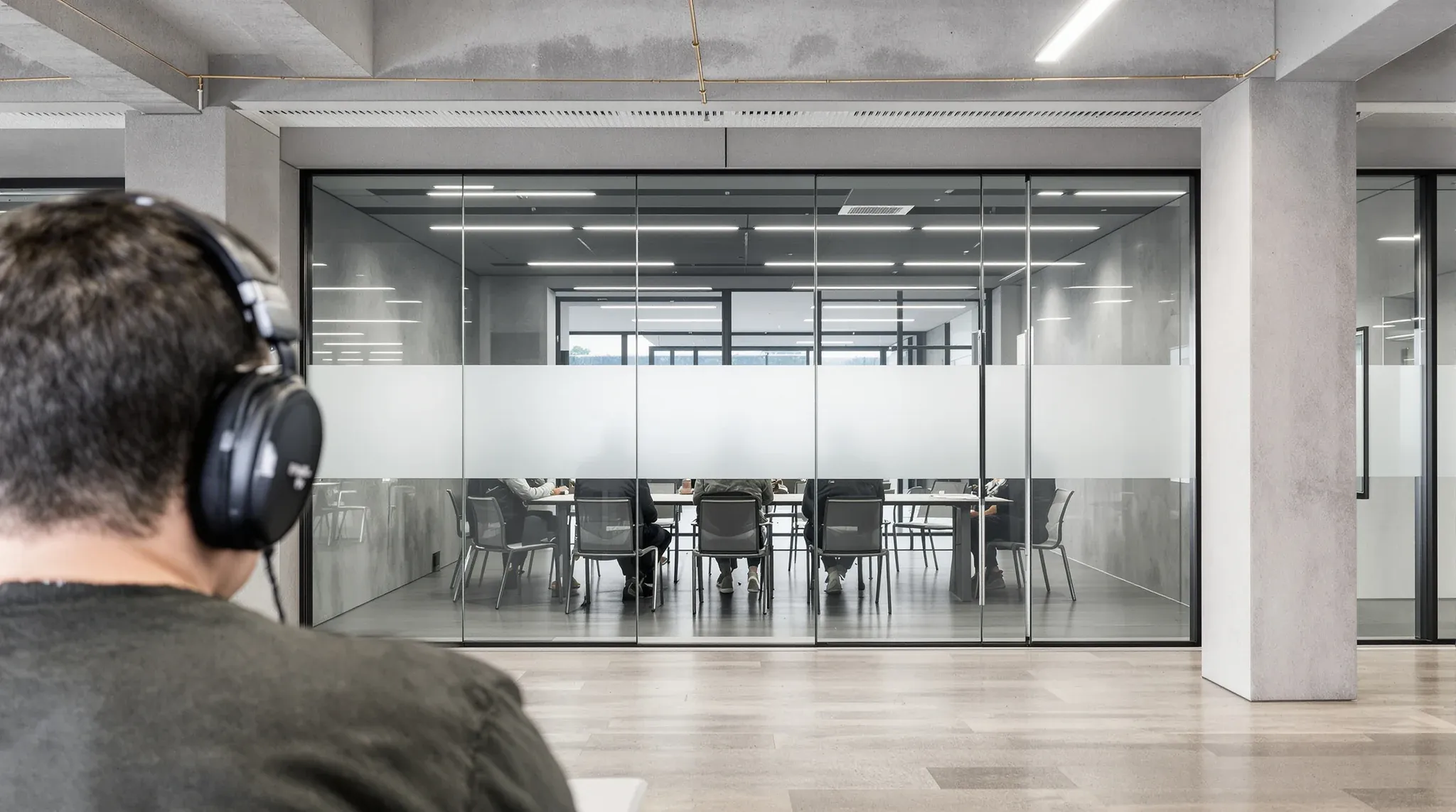 A person in headphones at a desk, seen from behind, facing a glass meeting room during a meeting