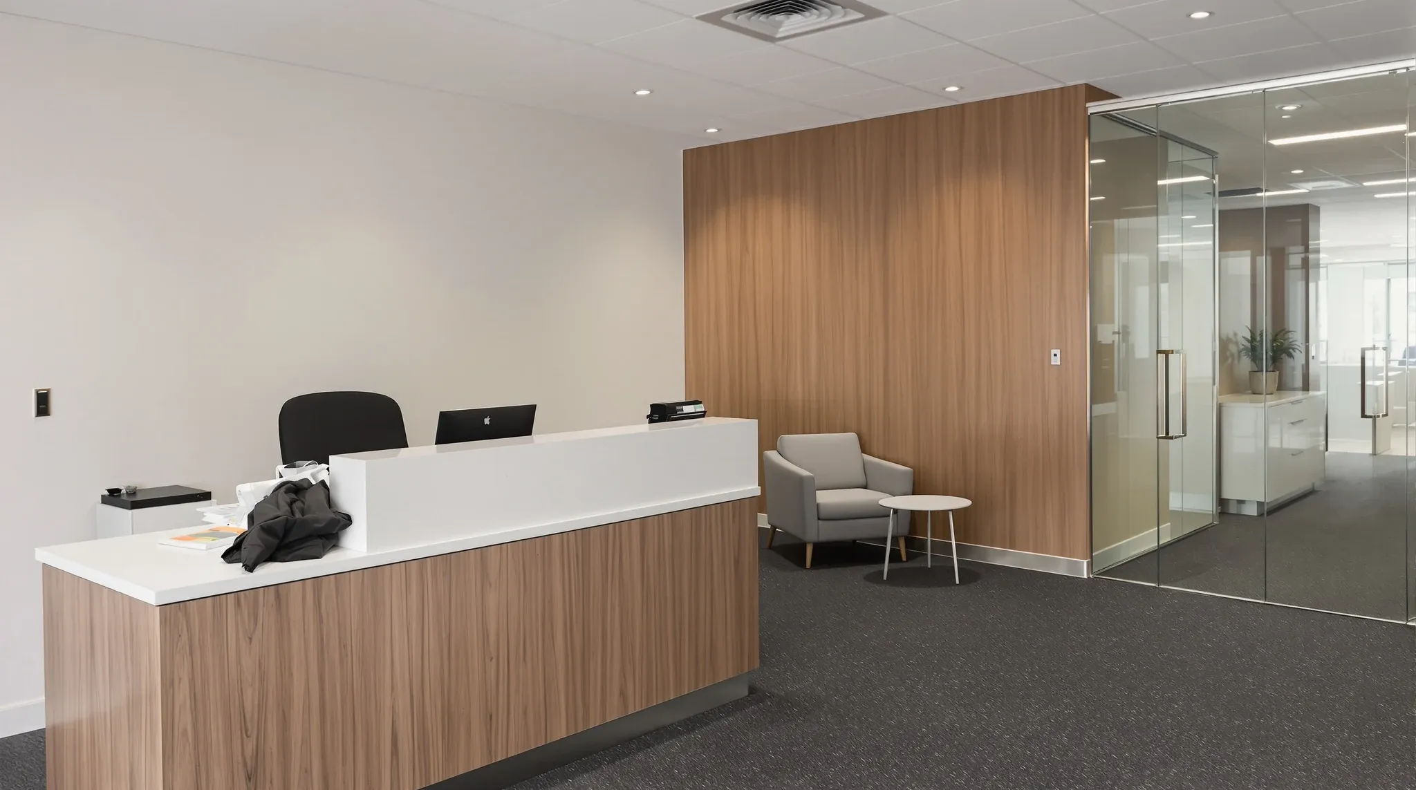 A law firm reception with a timber desk, two client chairs by a plain wall and a glass door to meeting rooms