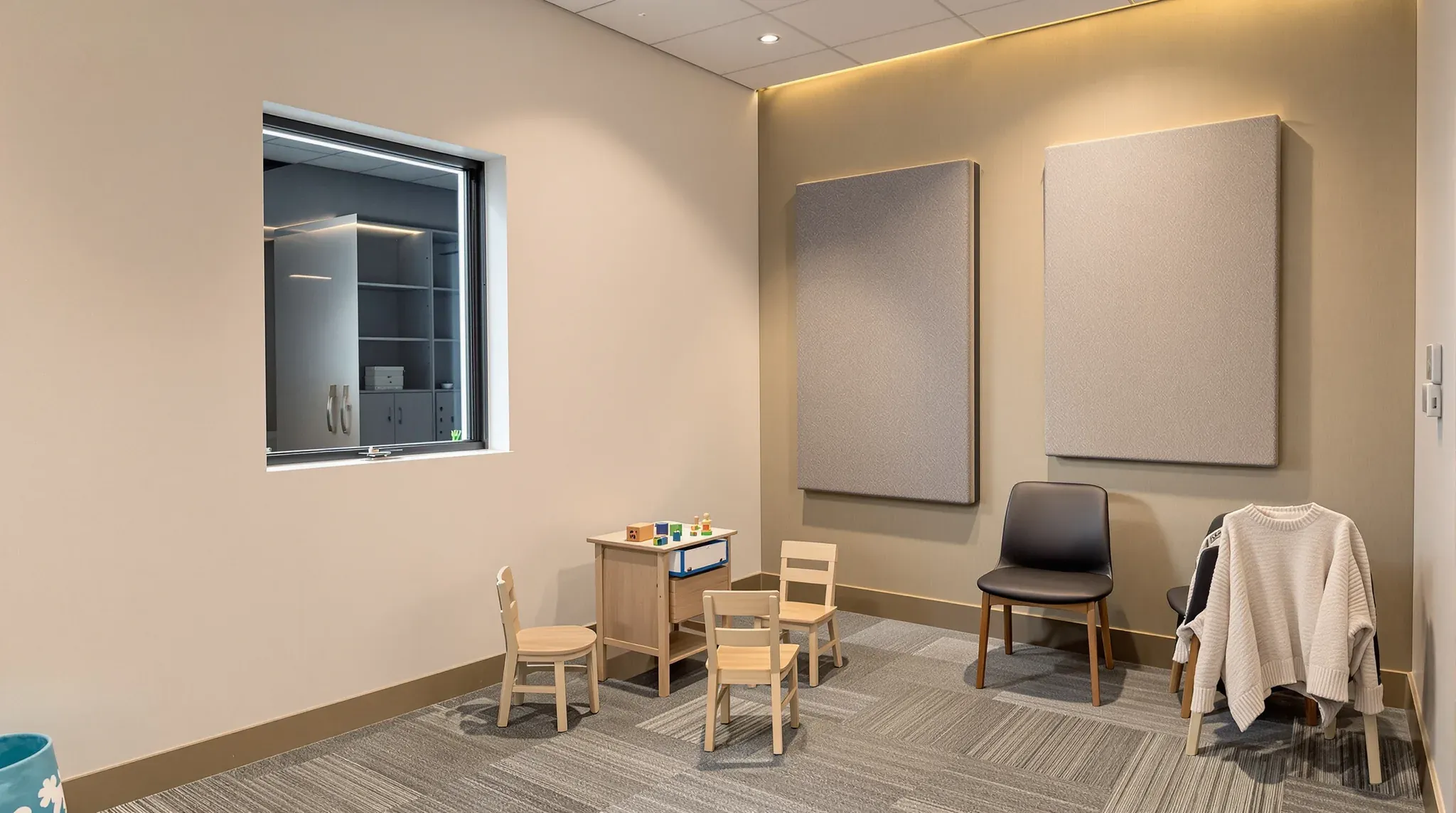 Small speech therapy room with child-size chairs, acoustic wall panels and an observation window into the next room