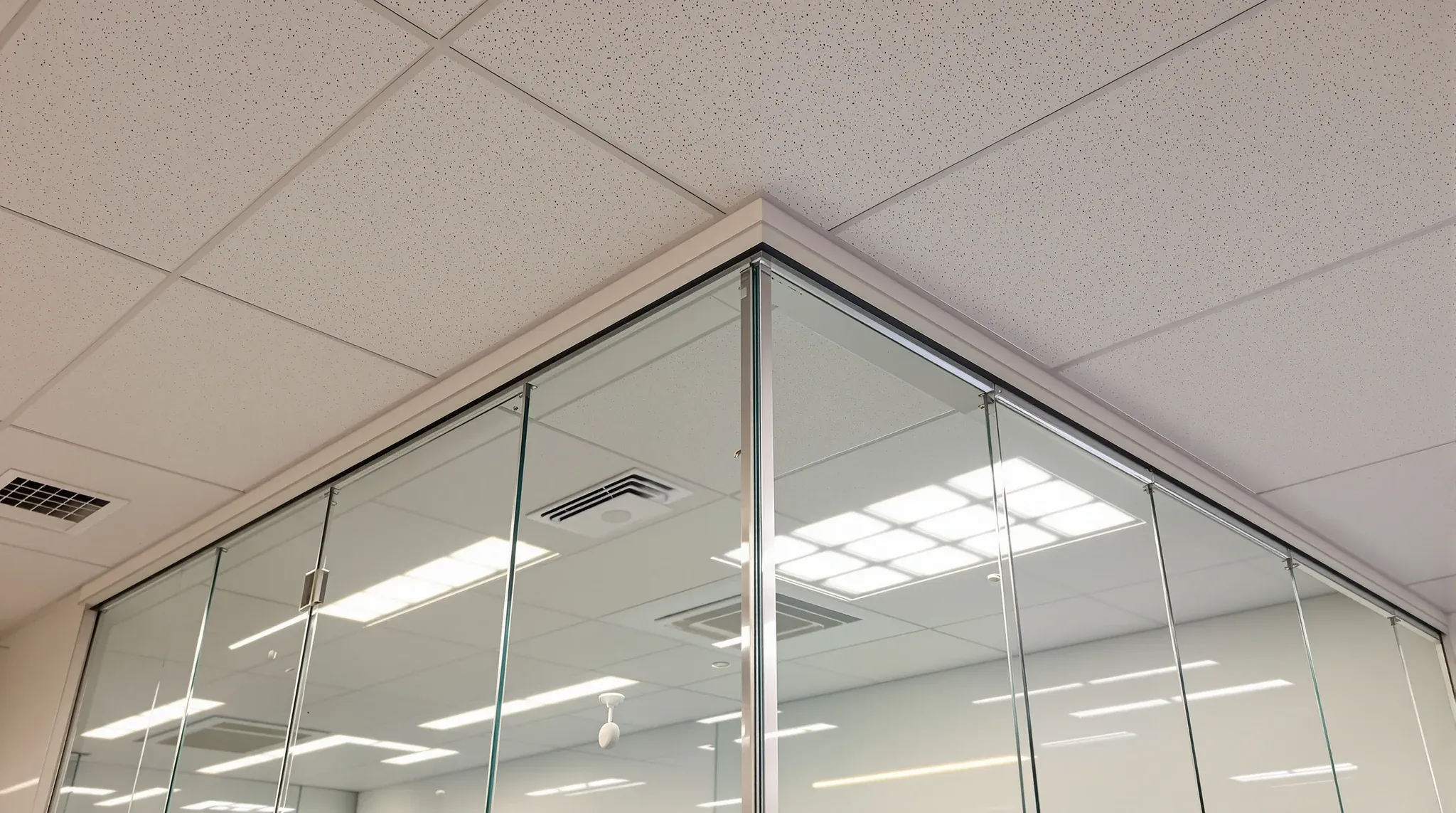 Looking up where a glass partition head lands exactly on a ceiling grid line, with lights and sprinklers aligned