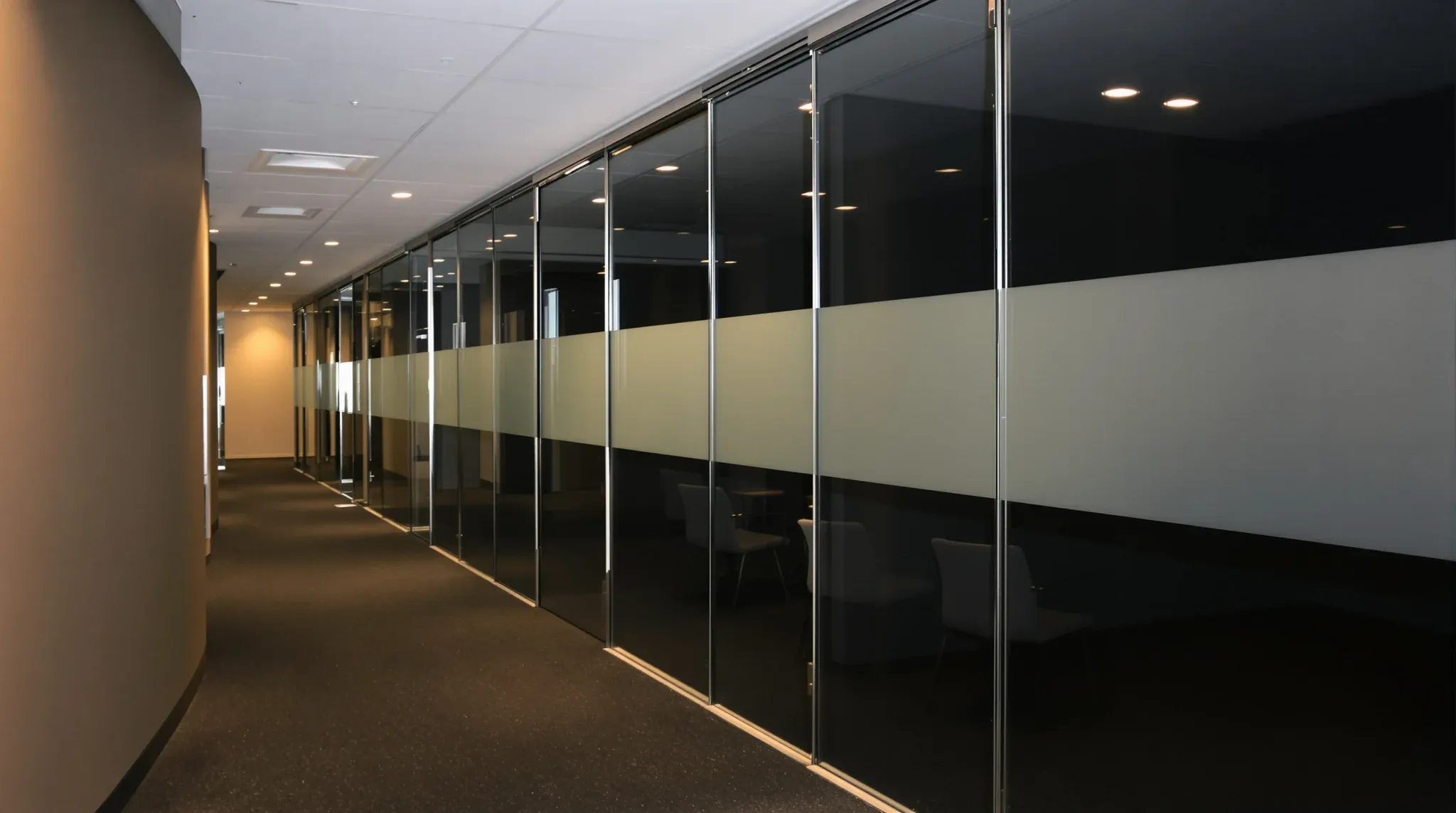 A long office corridor at night lined with equal frameless glass panels and dark rooms behind