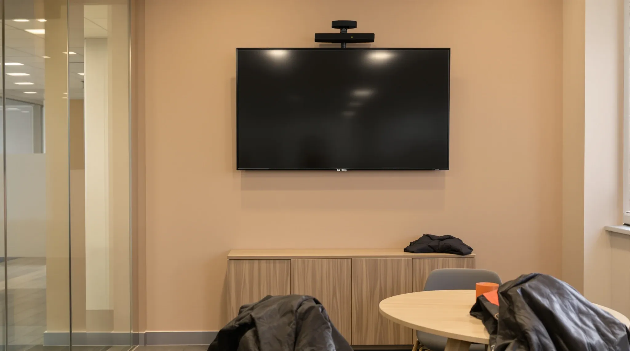 A small video call room with a large dark display mounted on a painted plasterboard wall above a low cabinet