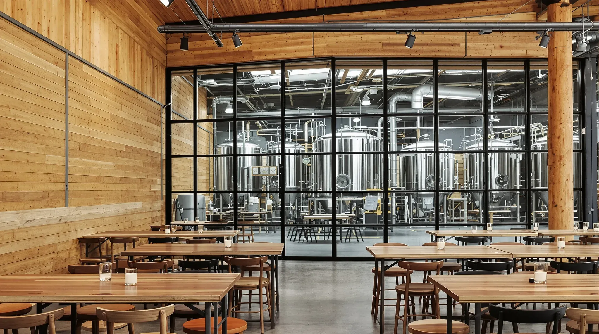Brewery taproom with long timber tables and a black steel-framed glass wall onto the stainless brewing tanks