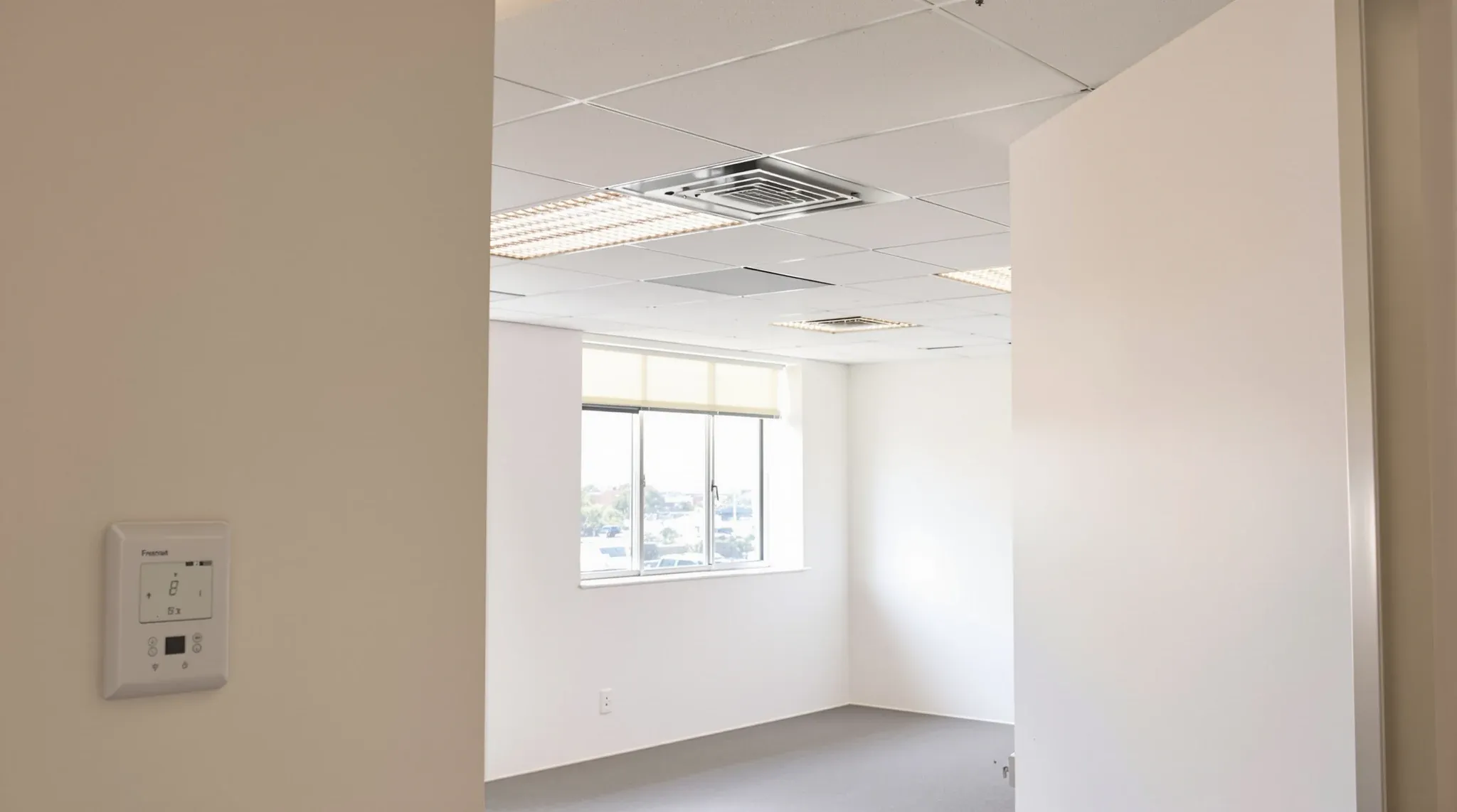Empty office with ceiling diffusers and a wall thermostat, window blinds half down