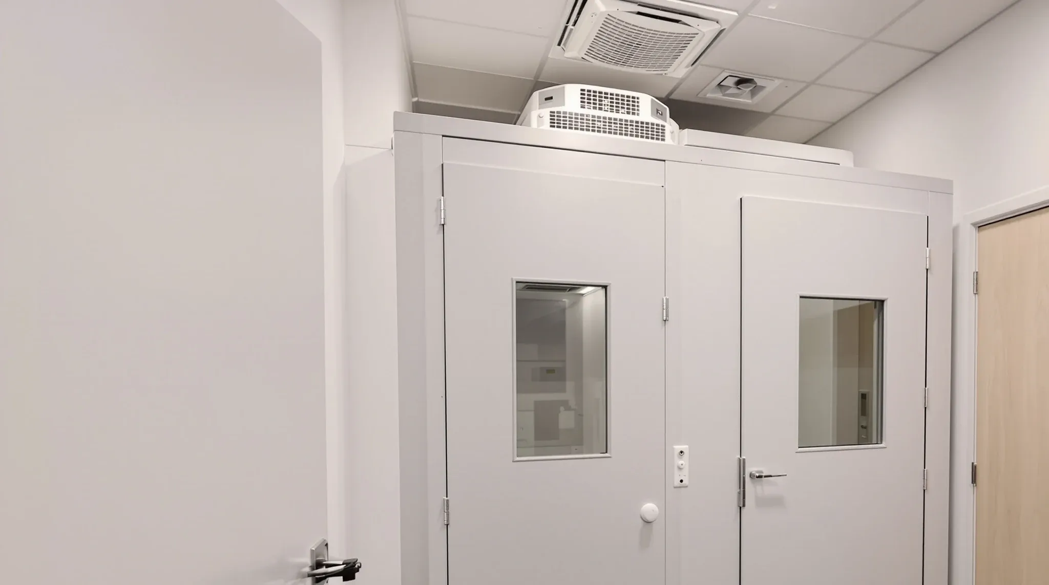 Prefabricated audiometric booth inside a plasterboard host room at a hearing clinic, with the clinician desk beside it