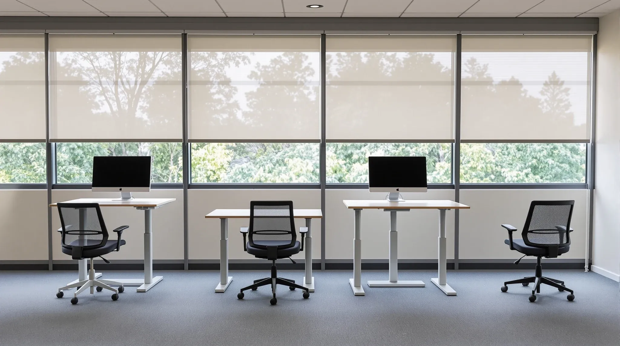 Four sit-stand desks facing half-lowered blinds in an office, one raised to standing height