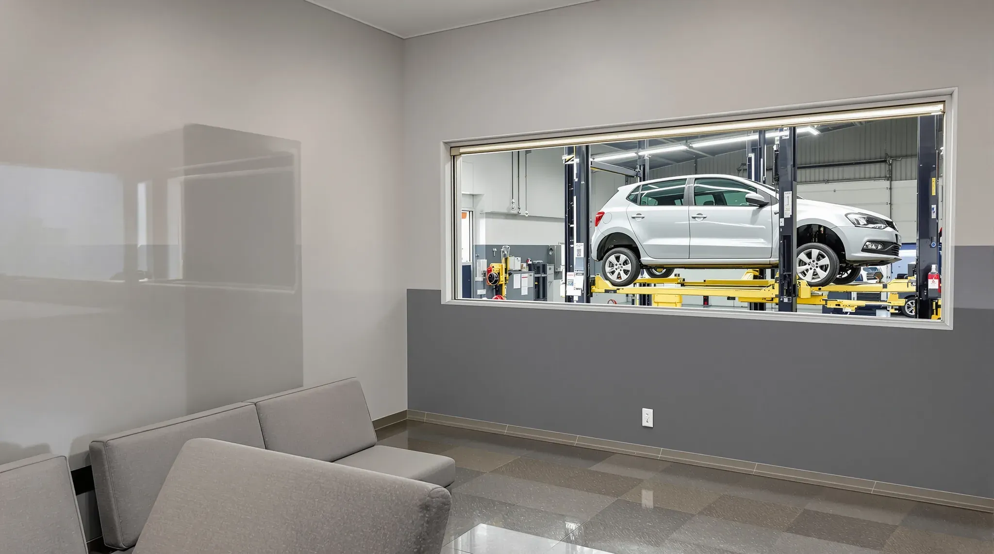 Tyre shop waiting room with grey seats and a long window into the workshop, where a car is raised on a hoist
