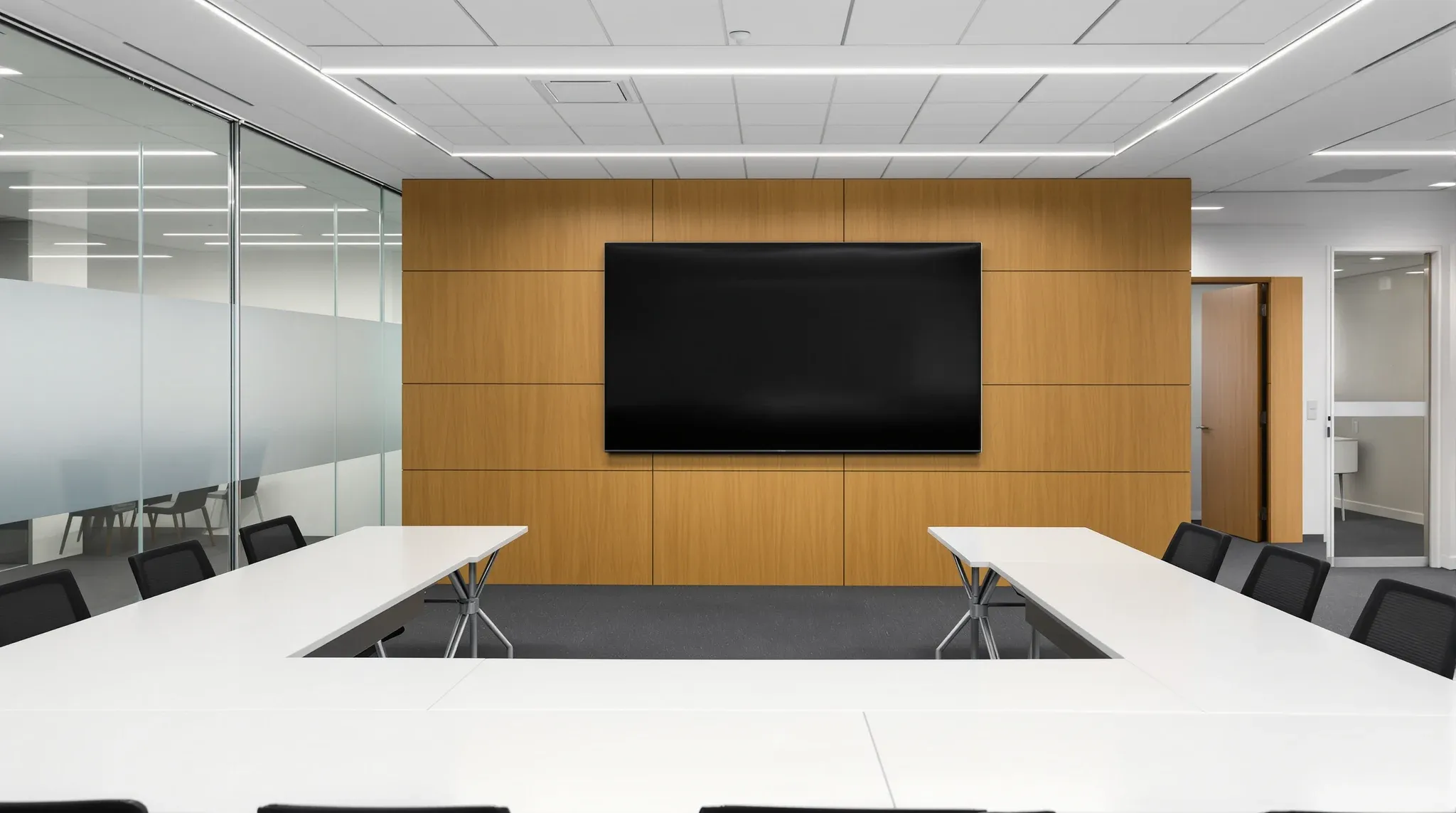 Empty corporate training room with U-shaped tables facing a blank wall screen and a glass wall to the corridor