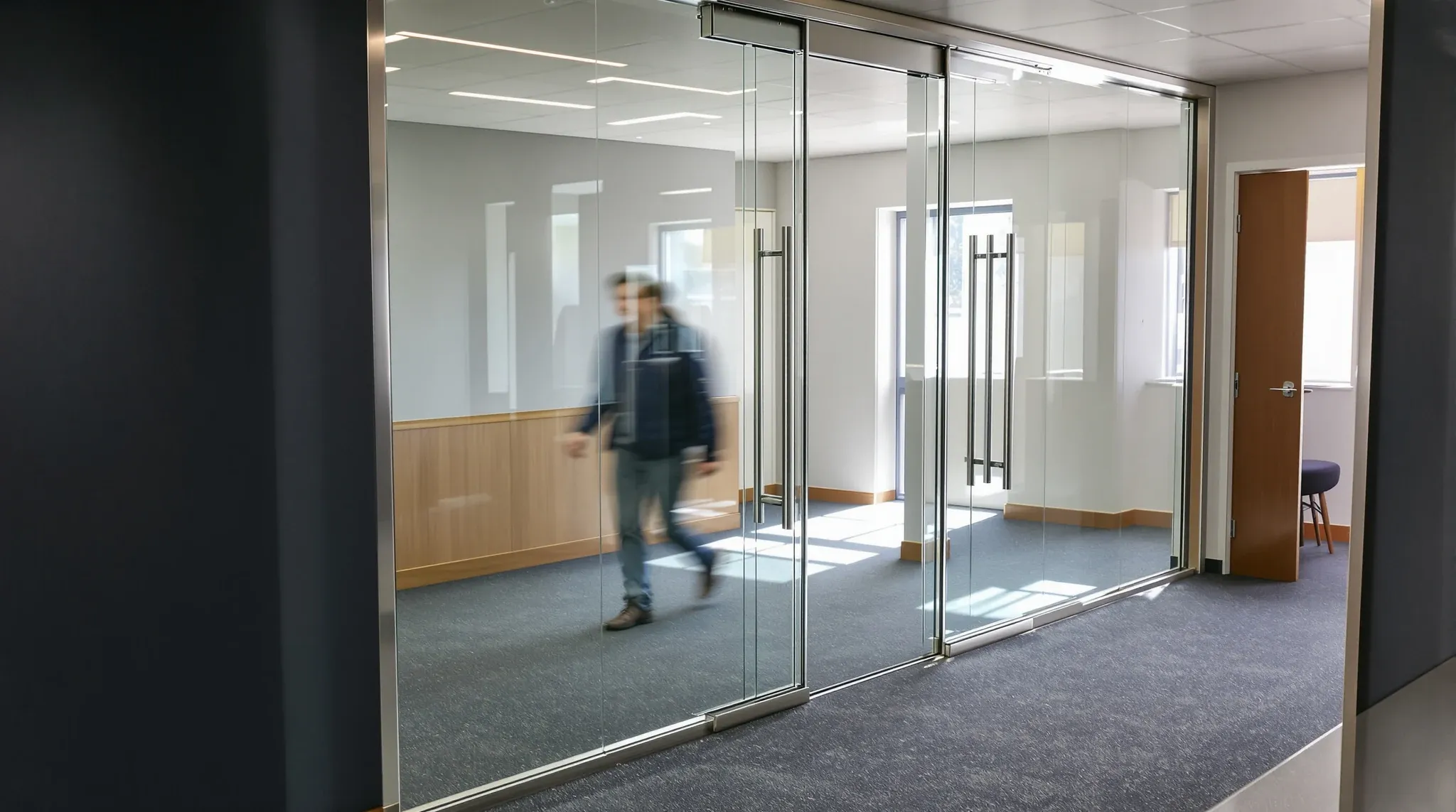 A tall frameless glass door reaching the ceiling in an office corridor, with a person walking through
