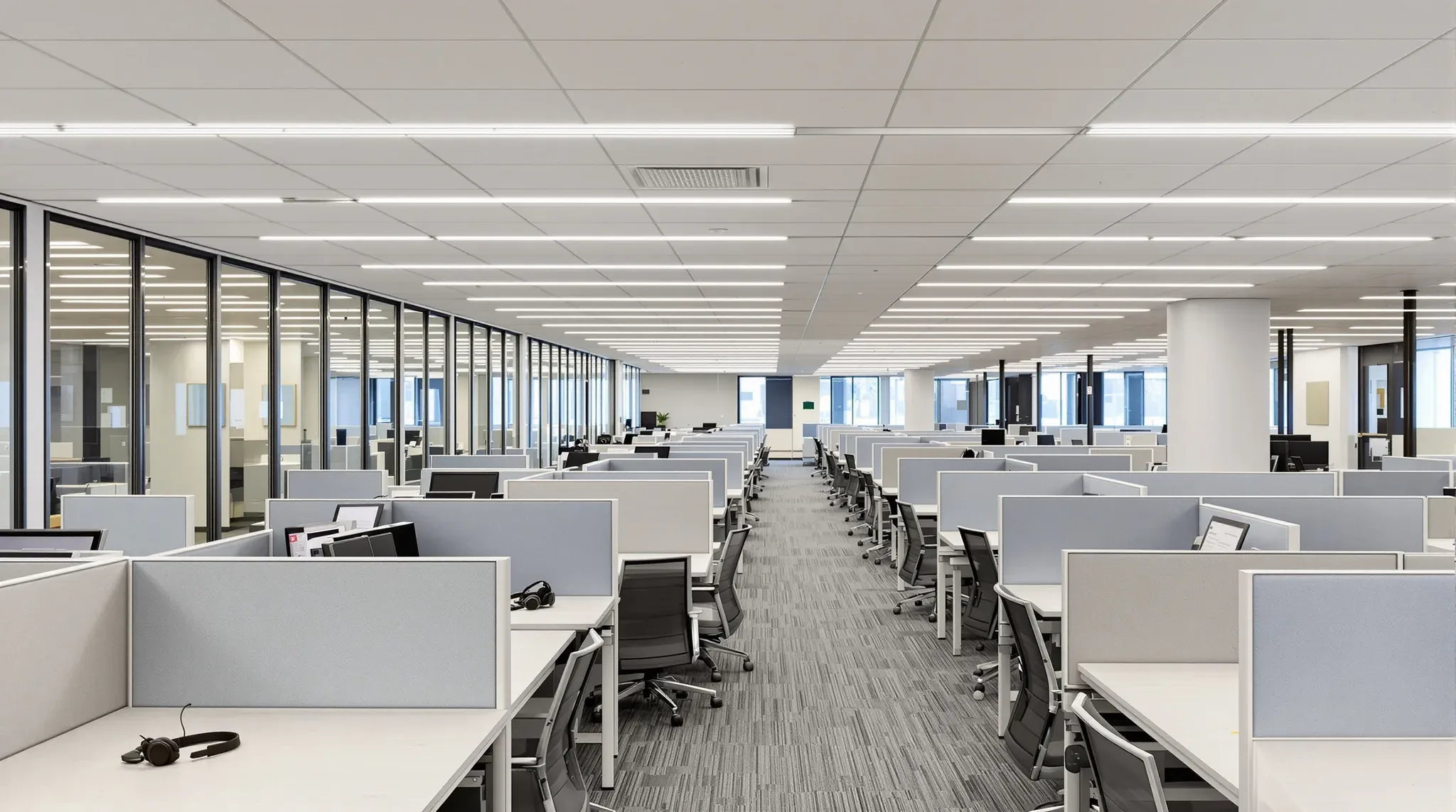 Contact centre floor with rows of desks behind low screens, a headset on a desk and glass meeting rooms