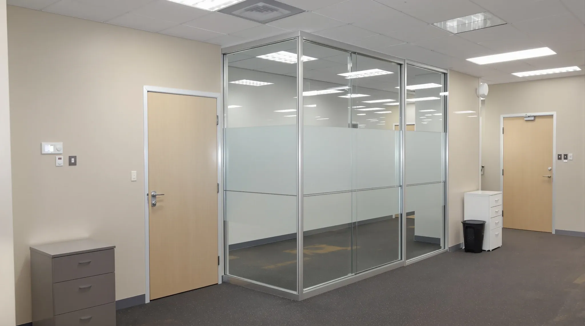 A plain painted office wall meeting a simple aluminium-framed glass partition under fluorescent light
