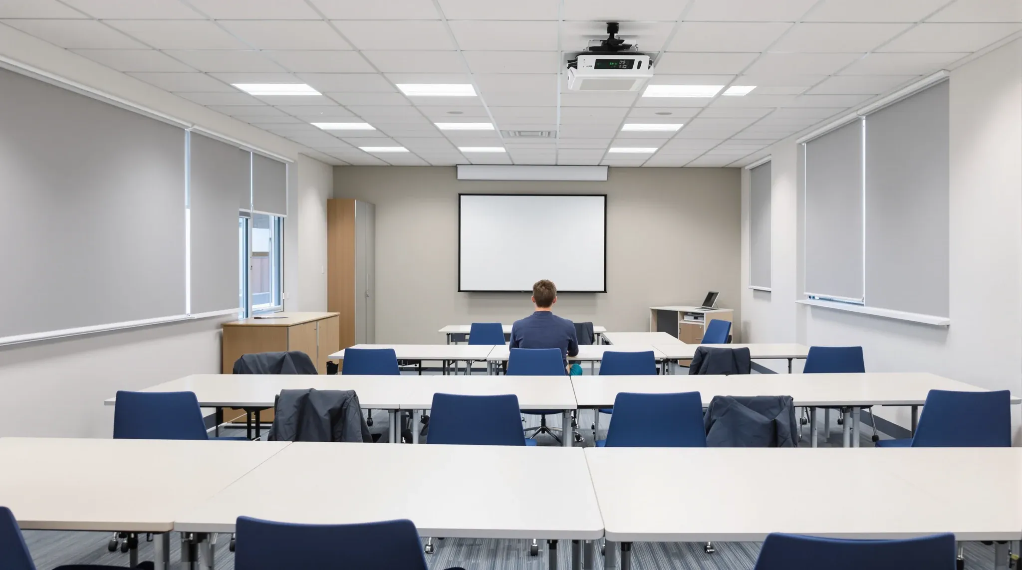 A training room with tables grouped for group work, a blank screen and one learner seen from behind