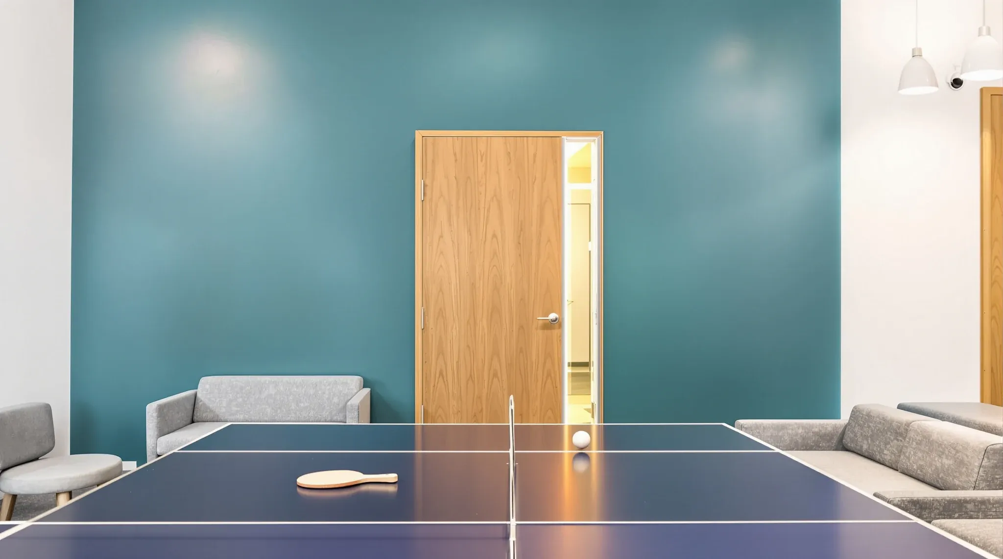 A table tennis table in an office breakout right beside the solid teal wall of a meeting room
