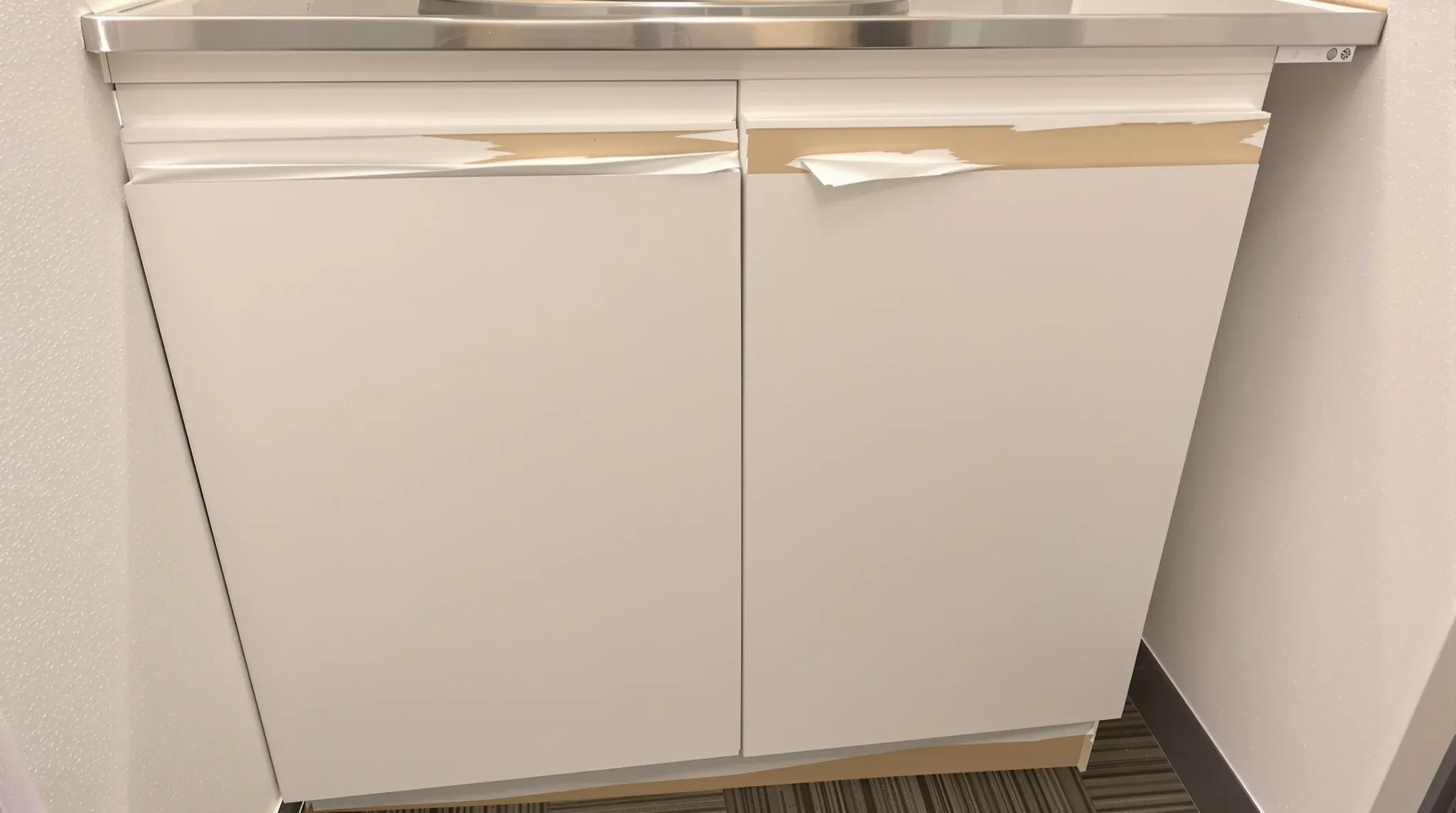 A swollen, peeling particleboard cupboard door under the sink in a small office kitchenette