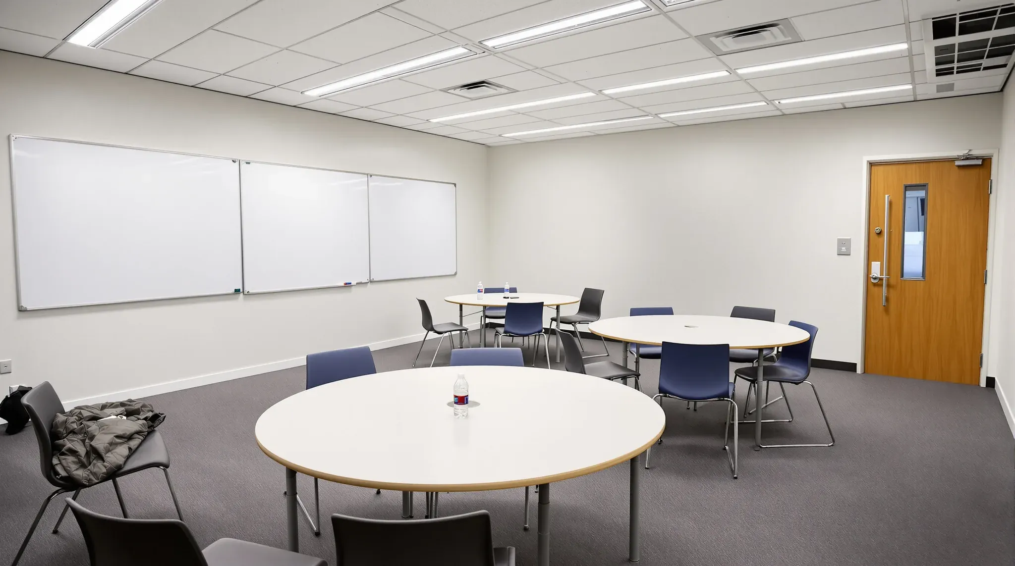 University teaching room with round group tables, whiteboards along the side wall and a door with a vision panel