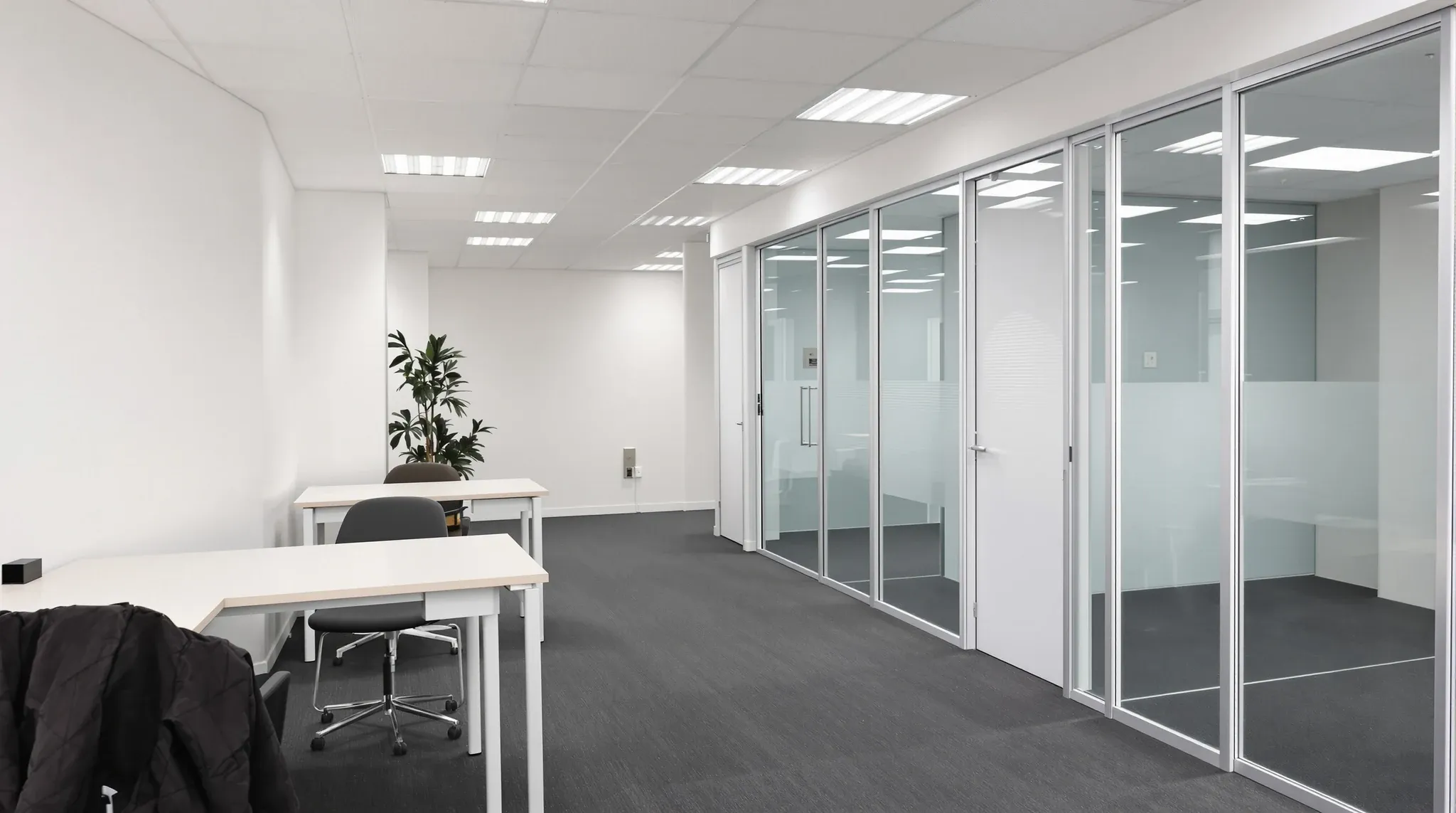 A small carpeted office with a plasterboard storeroom and two glass-fronted rooms beside two rows of desks