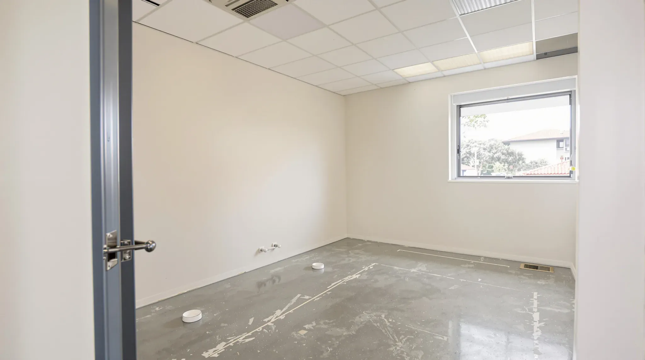 Empty former consulting room after make good, with capped pipes low on the wall, a bare floor and a window
