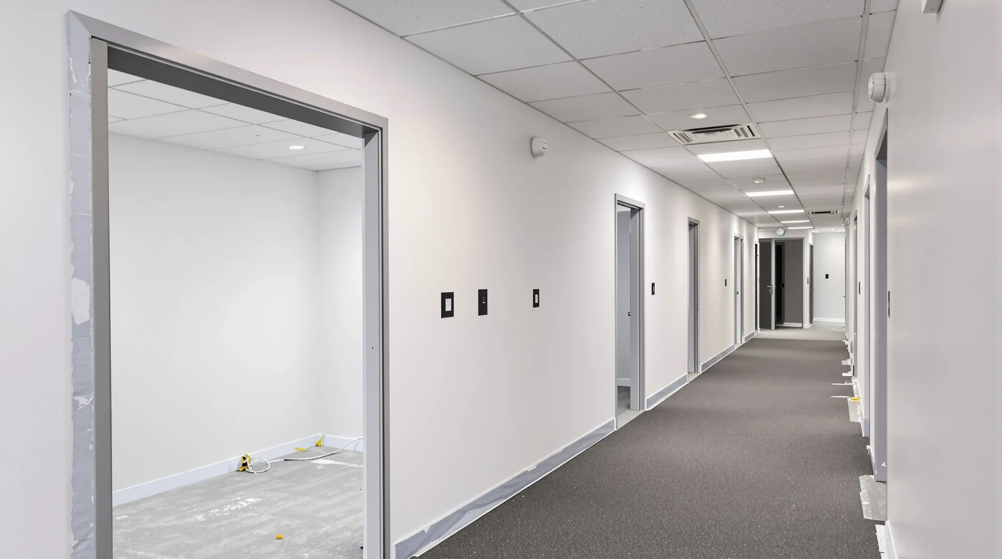 A near-finished office corridor with fire doors held open and sprinklers and lights aligned in the ceiling