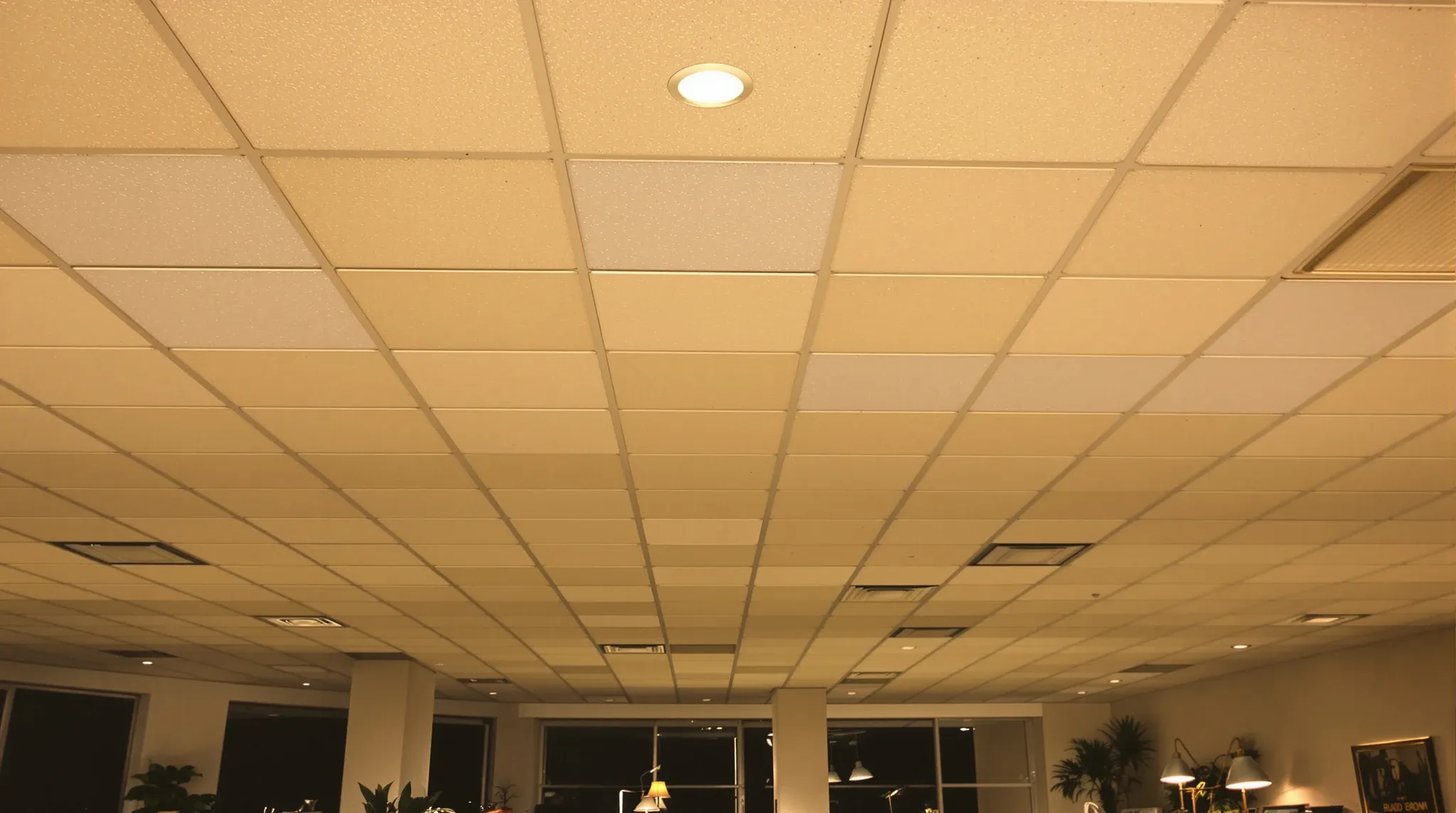 An older office ceiling of tired, yellowed and slightly sagging tiles above desks still in use