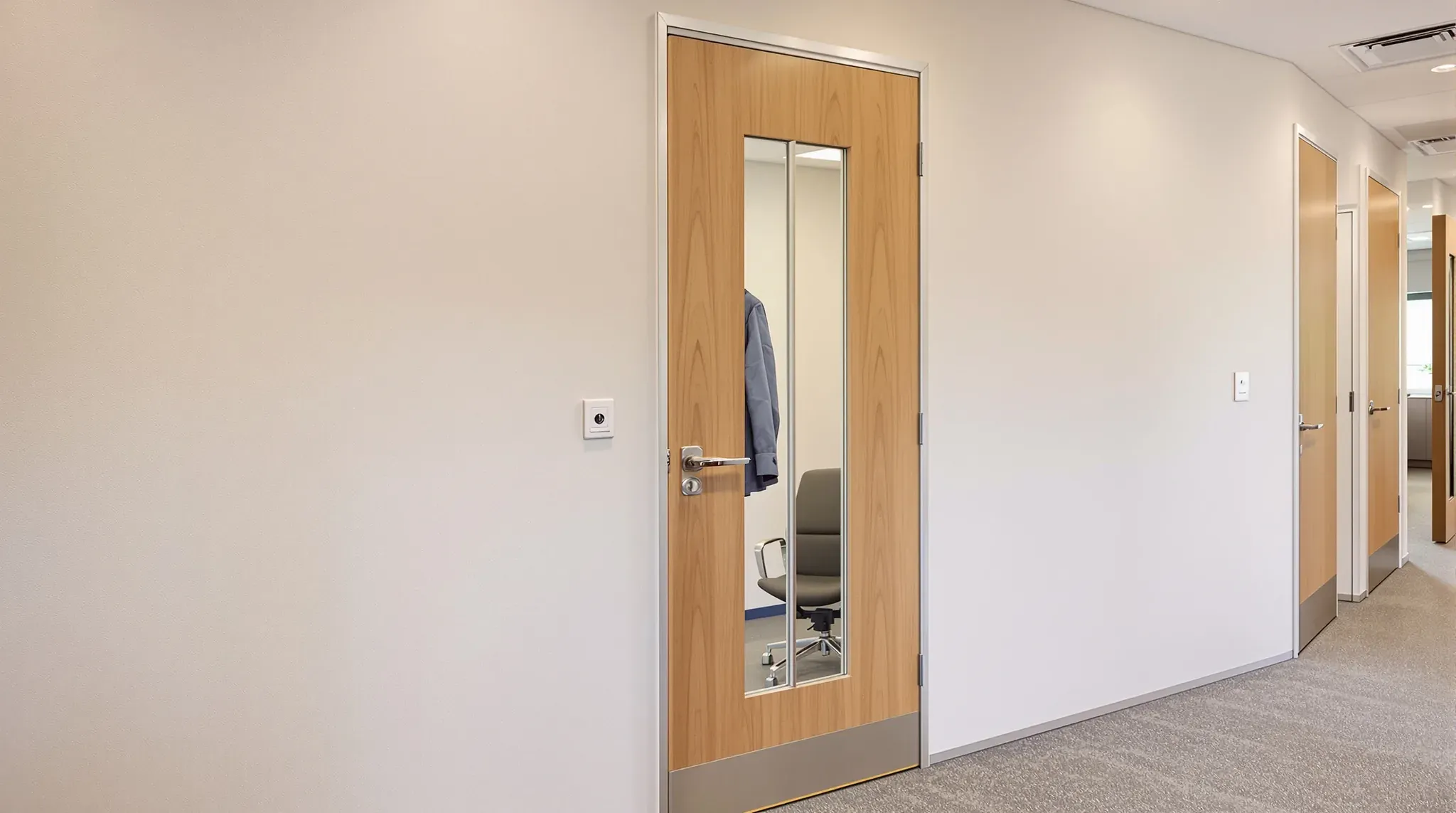 Timber meeting room door with a narrow vision panel in a plasterboard corridor wall, a jacket visible inside