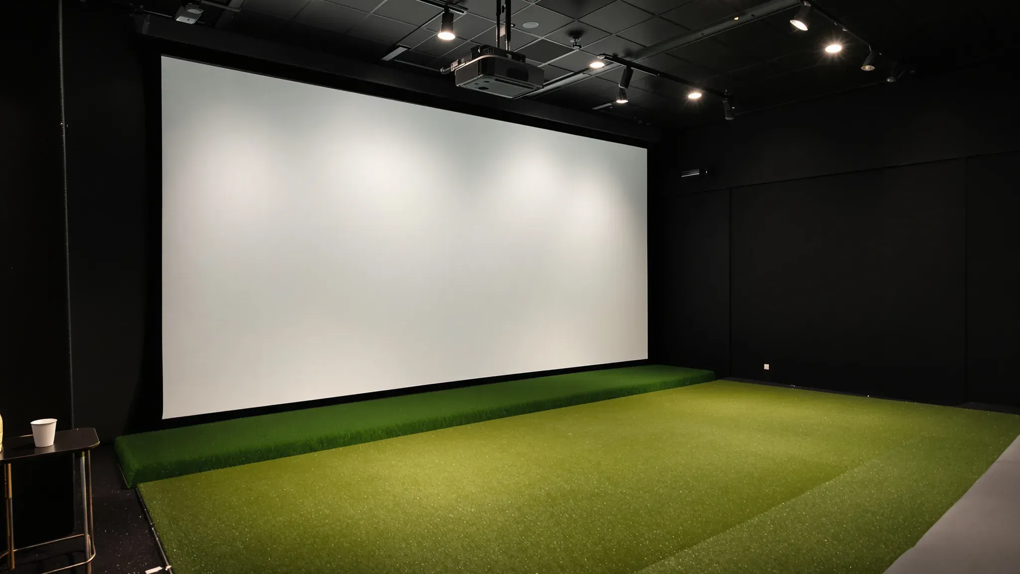 Golf simulator bay with a large white impact screen, green hitting turf, black walls and a ceiling projector