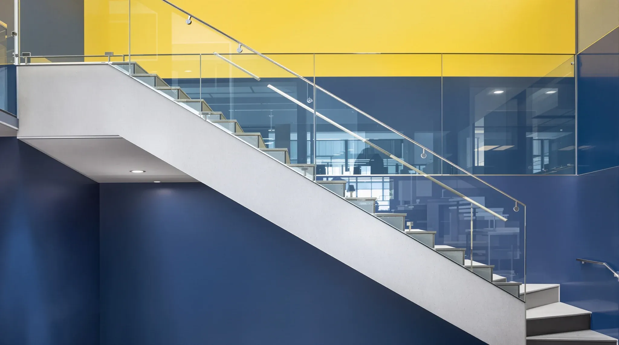 A straight office stair with a glass balustrade, the lower floor painted blue and the upper floor yellow