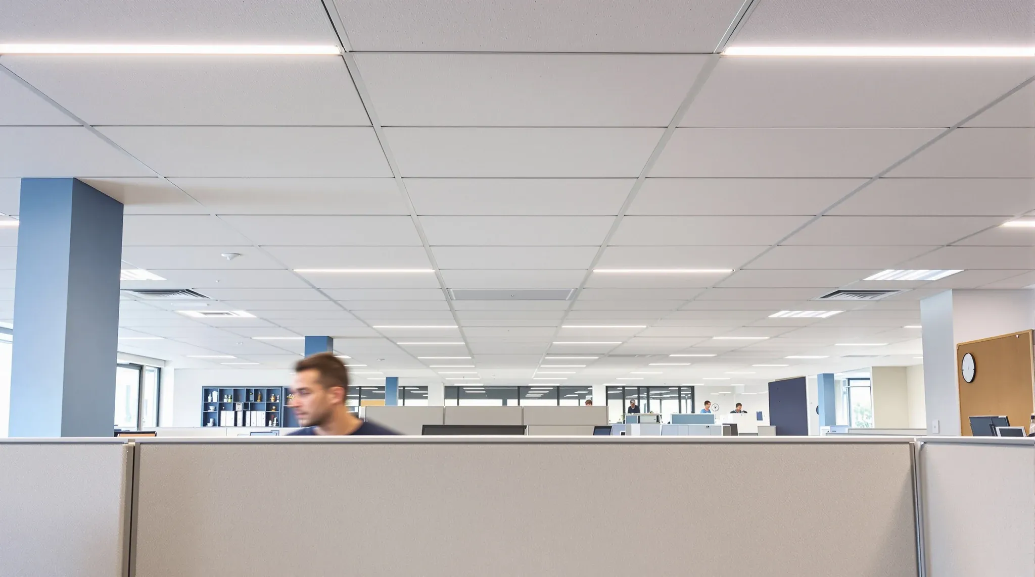 A new acoustic ceiling with slim LED lights installed above the existing, older desks in an office