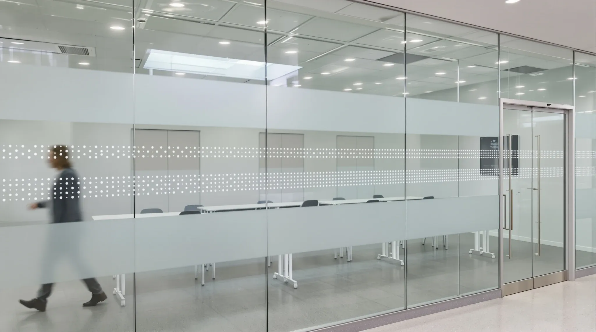 A full-height glass wall with dotted frosted safety bands and a person blurred walking past