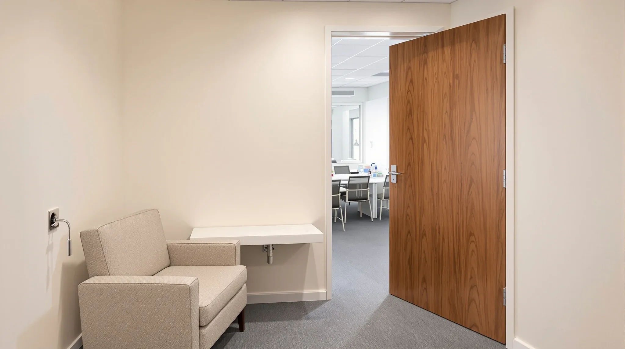 A small parents room with a nursing armchair and a hand basin, its single door ajar to the office